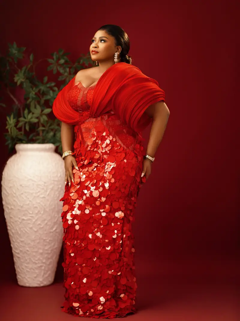 A woman in a red sequinned Ablizz gown with sculpted pleated sleeves, against a red backdrop